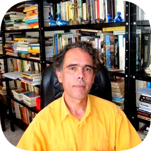 ​​​​Prof. Dr. Henrique Carneiro, de camisa amarela, sentado diante de uma estante com livros